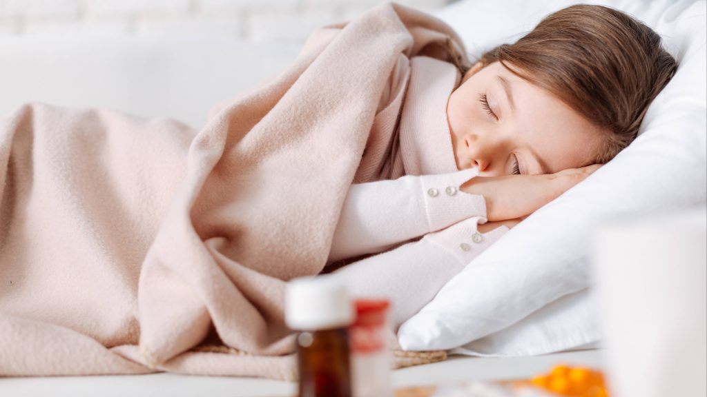 a little white school aged girl asleep in bed, covered in a blanket with medicine on a side table nearby, perhaps because she's sick and not feeling well with a cold or fever