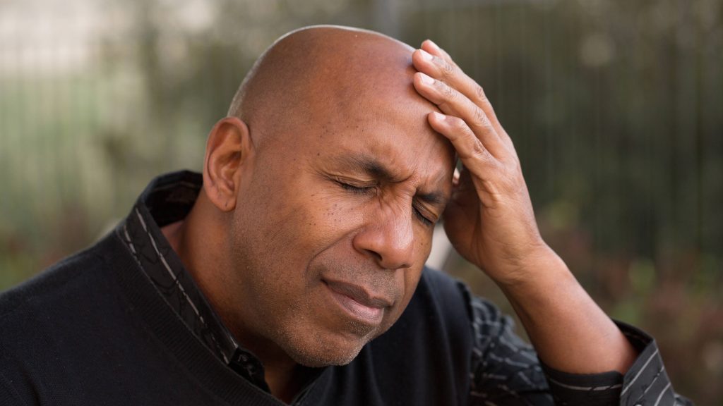 a middle-aged Black man sitting outside, with his eyes closed and resting his head in his hand as if he's in pain, with a tension headache