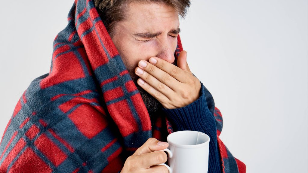 a young white man looking sick or cold, wrapped in a plaid blanket, holding a mug with tea and getting ready to sneeze or cough into his hand