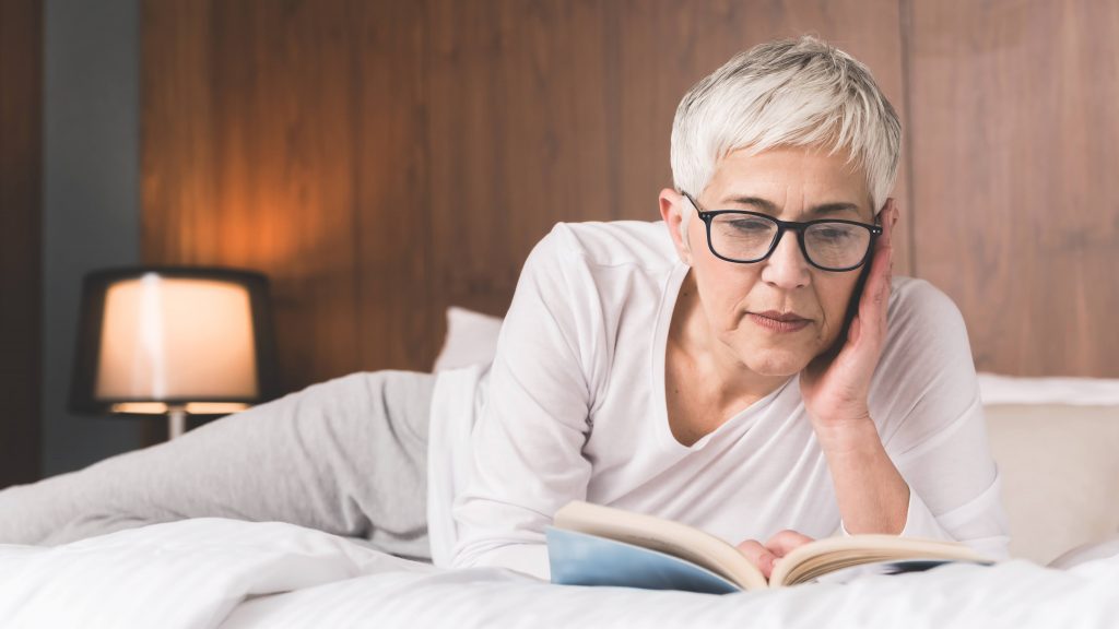a white middle aged woman with light gray hair, wearing glasses and resting, relaxing a bed reading a book
