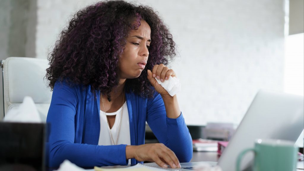 a young Black woman looking sick or ill working at a computer on a table and holding a tissue to her face as she coughs or sneezes
