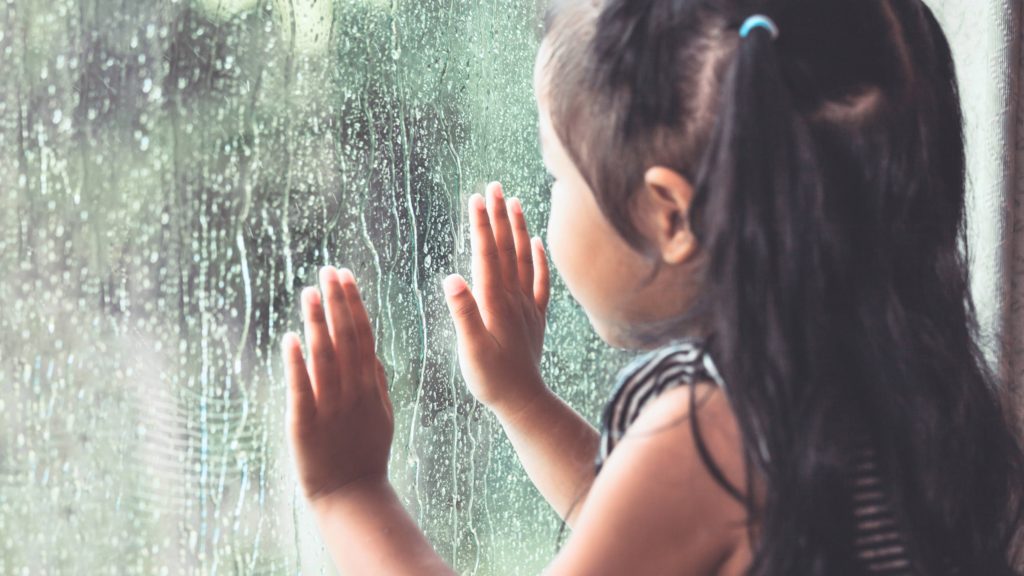 a young school aged perhaps Latina or Asian girl with ponytails, during a storm looking out a window with raindrops falling on the pane