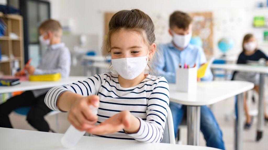 a young white girl in a school classroom, wearing a mask and putting sanitizer on her hands while sitting at her desk, social distancing from classmates