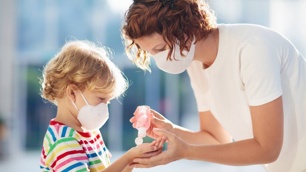 an adult white woman wearing a mask and putting hand sanitizer in the hands of a young white child who is also wearing a mask