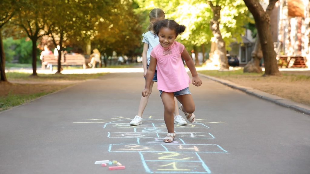 two little girls, one Black and one caucasian, playing hopscotch outside in a park with trees on a sunny day