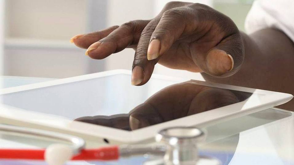 closeup of a Black person, perhaps in a medical office, with his or her hand on a iPad tablet with a stethoscope next to it on a table (original)