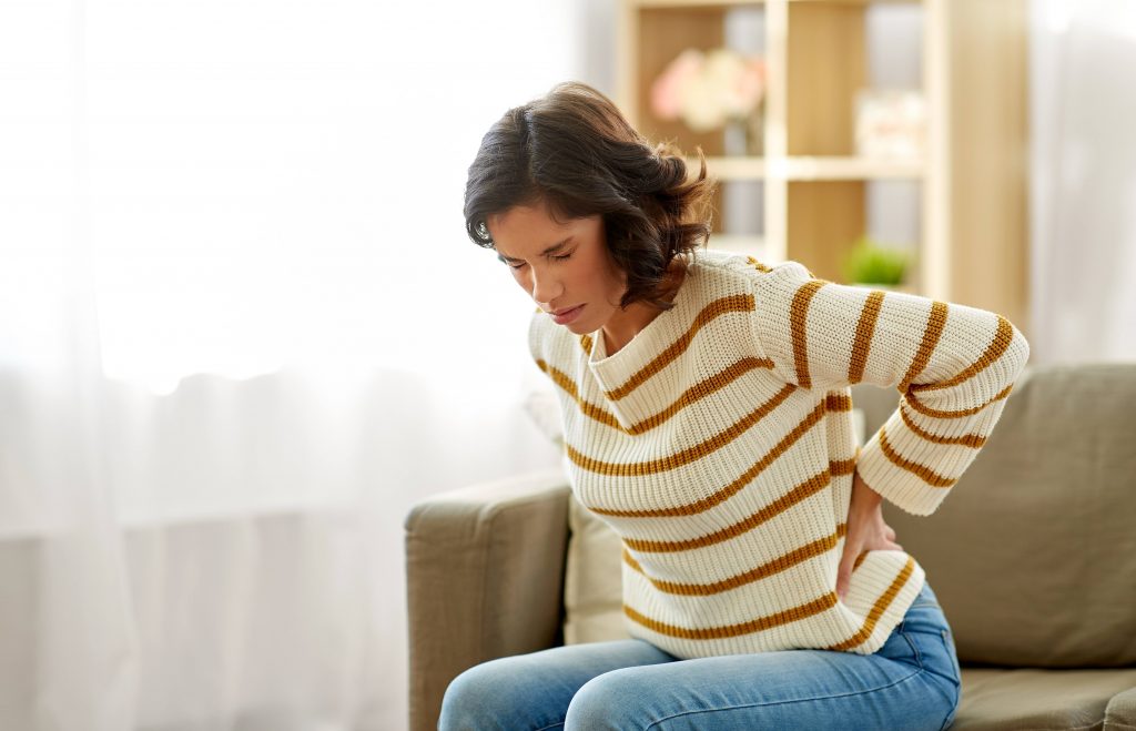 a young white woman in a striped sweater, sitting on a couch, wincing in pain, her eyes closed, looking sad and holding her back because it hurts