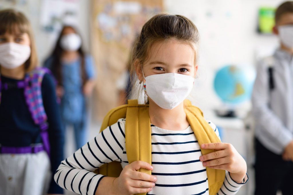 a young white schoolgirl in a classroom wearing a backpack, with other students all wearing masks and social distancing
