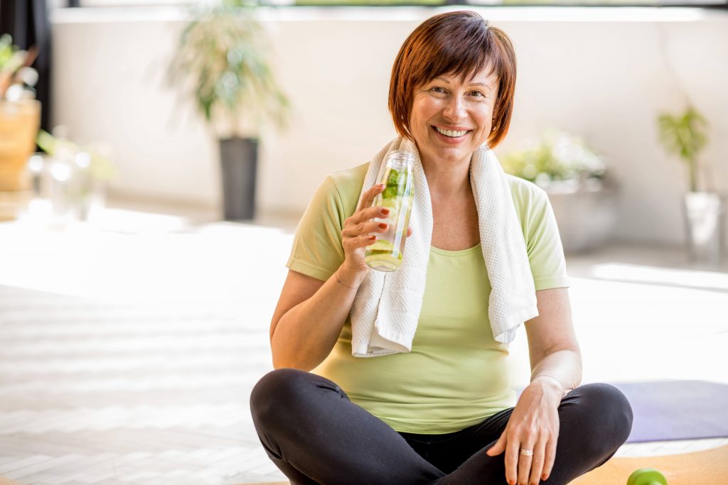 a white or perhaps Latina middle aged woman smiling and sitting on the floor with a towel and a bottle of water after exercising