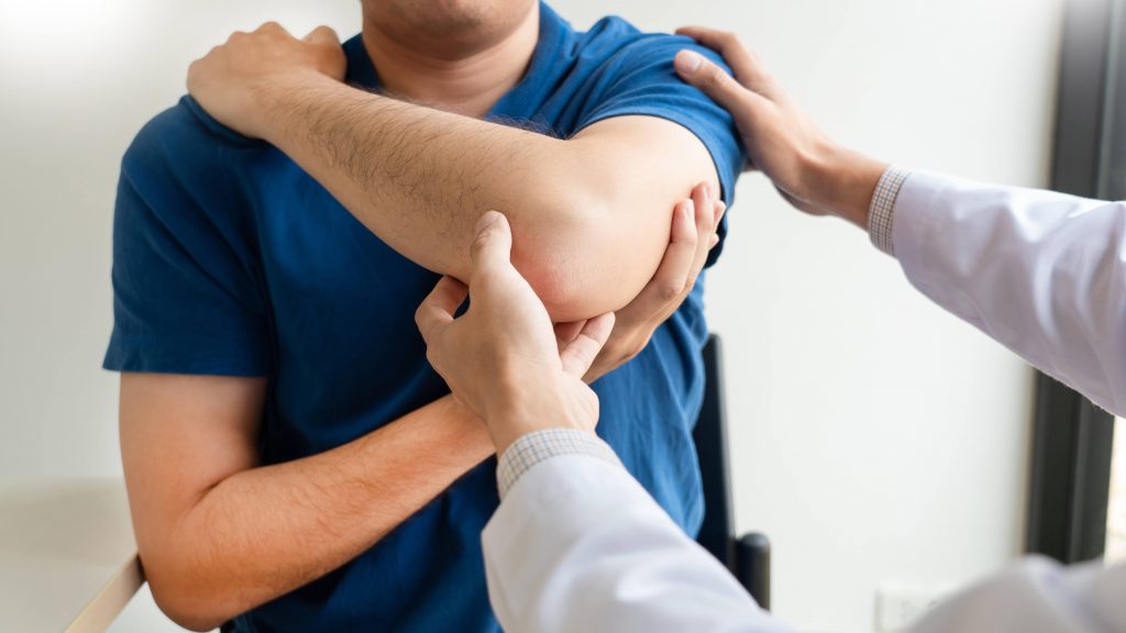 Un hombre con camisa azul levanta su brazo izquierdo adolorido, posiblemente debido a una lesión en el hombro o el codo, mientras un profesional médico examina el problema. 