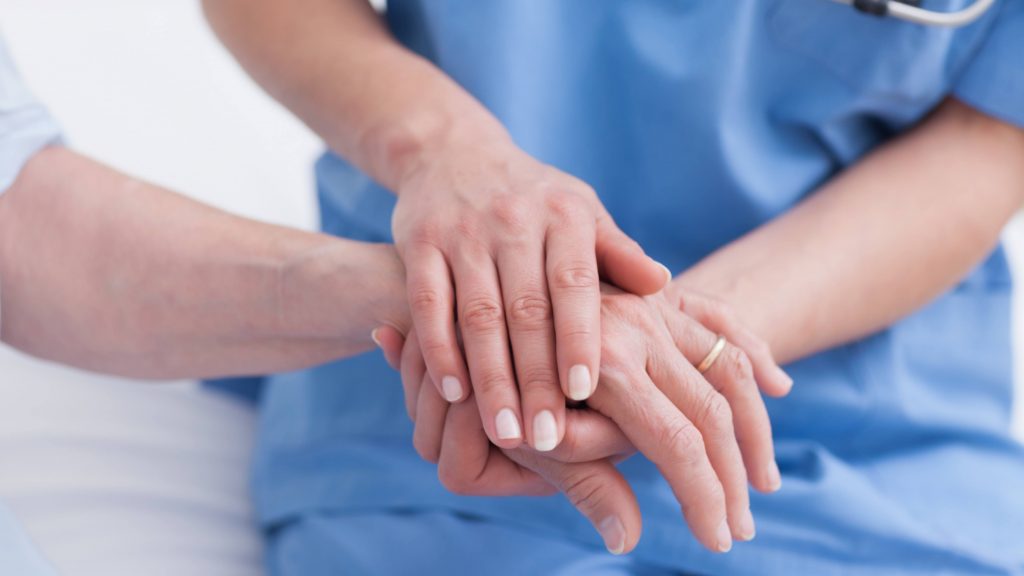 a white medical staff person in blue scrubs holding the hand of a white patient showing concern and care