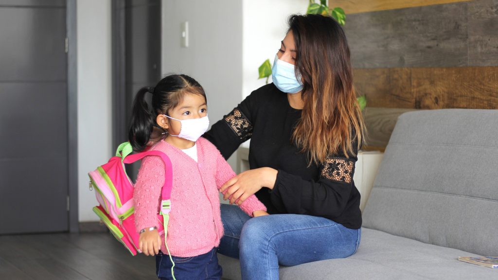 a young, perhaps Latina woman, sitting on a couch helping a little Latina girl, maybe her daughter, get ready for school a adjusting her backpack and both wearing face masks