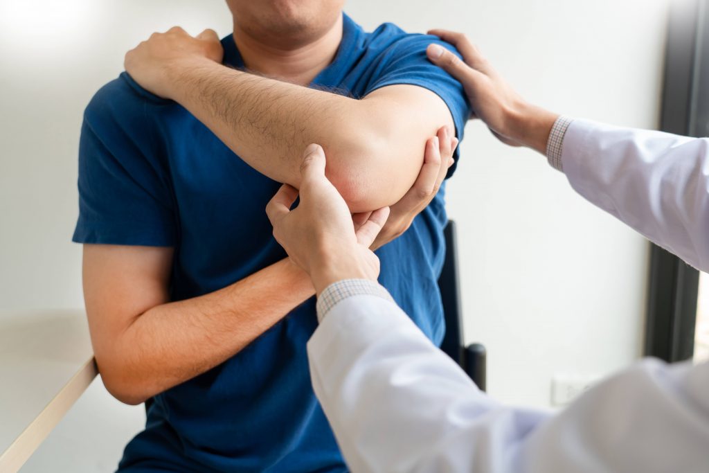 a white man in a blue shirt holding up his sore left arm, in pain, perhaps an injured shoulder or elbow, with a medical person examining the injury