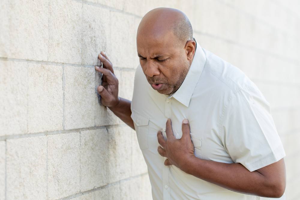 a Black middle aged man in a white shirt holding his hand to his chest and leaning against a wall in pain, short of breath and perhaps having a heart attack