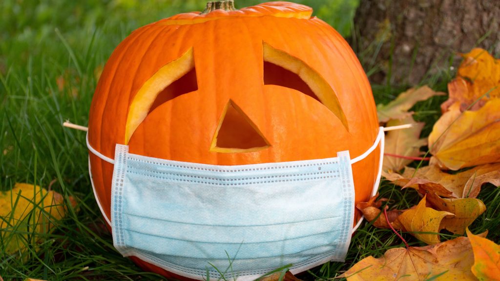 a carved Halloween pumpkin sitting out in the grass and leaves, wearing a PPE face mask