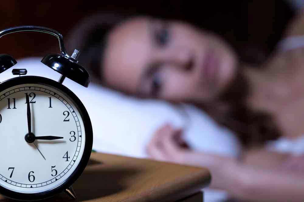 a close-up of an alarm clock reading 3 o'clock, with a wide awake woman lying on a pillow in the background, out of focus, trying to sleep and suffering from insomnia