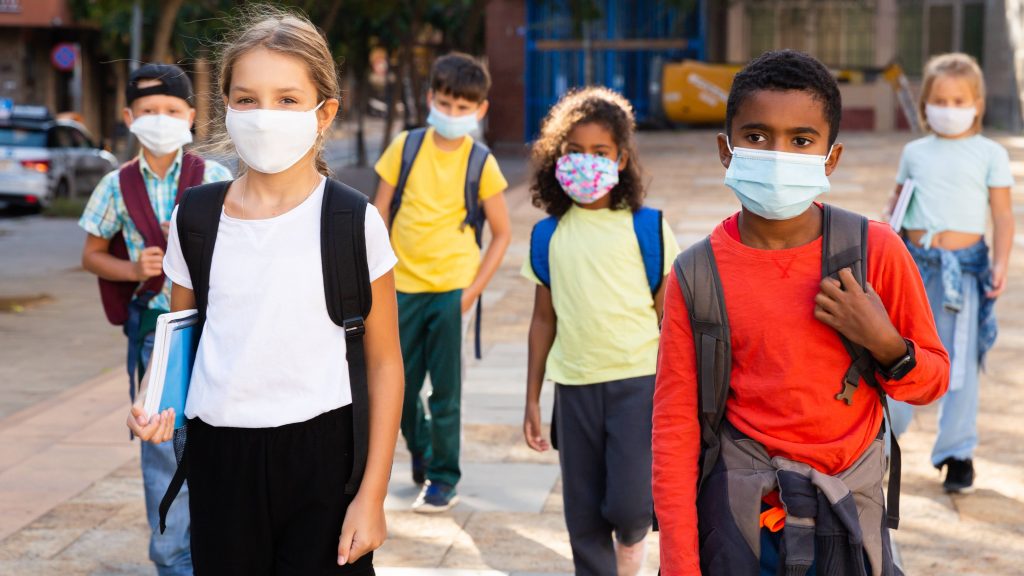 a diverse group of school aged children, tweens, wearing face masks and carrying backpacks, walking together with several feet between them