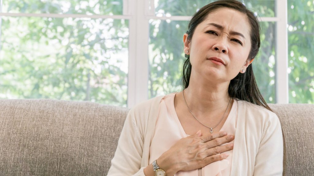 a middle aged Asian woman sitting on a couch near a window with her hand on her chest looking worried, sad, perhaps in pain