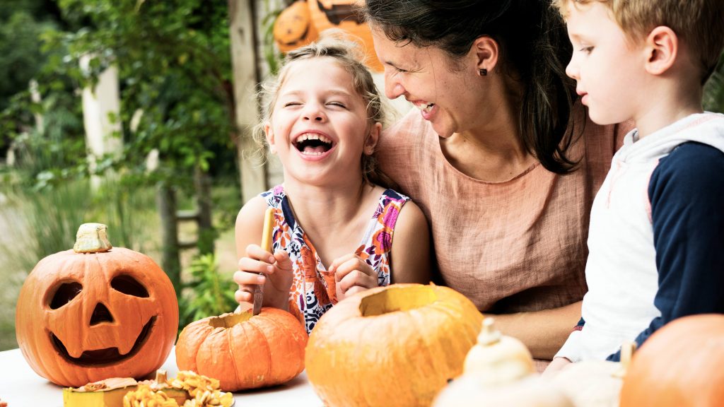 a white adult woman, maybe a mom, laughing and smilng with two white children as they make Halloween arts and craft activities