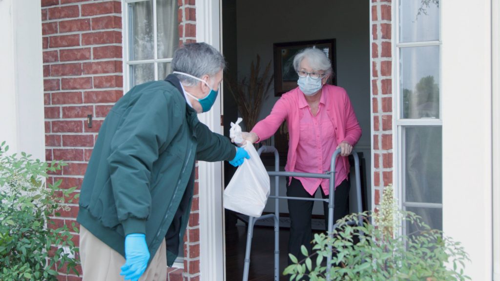 a white middle aged man wearing a mask and gloves delievering food to an older white woman, also wearing a mask, at her front door during COVID-19 pandemic