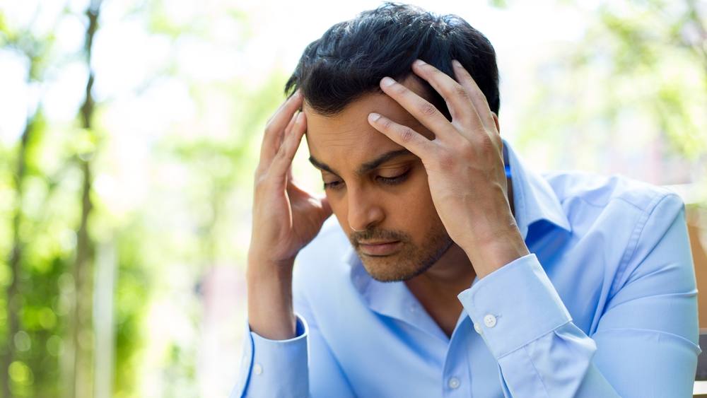 a young adult man, perhaps Latino, wearing a blue shirt and holding his head in his hands looking sad, stressed, depressed, unhappy