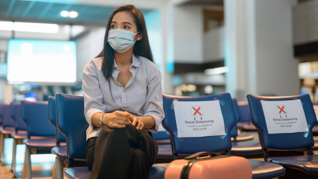 a young adult woman, perhaps Asian or Latina, sitting in an airport with a suitcase and social distancing COVID-19 signs on the seats