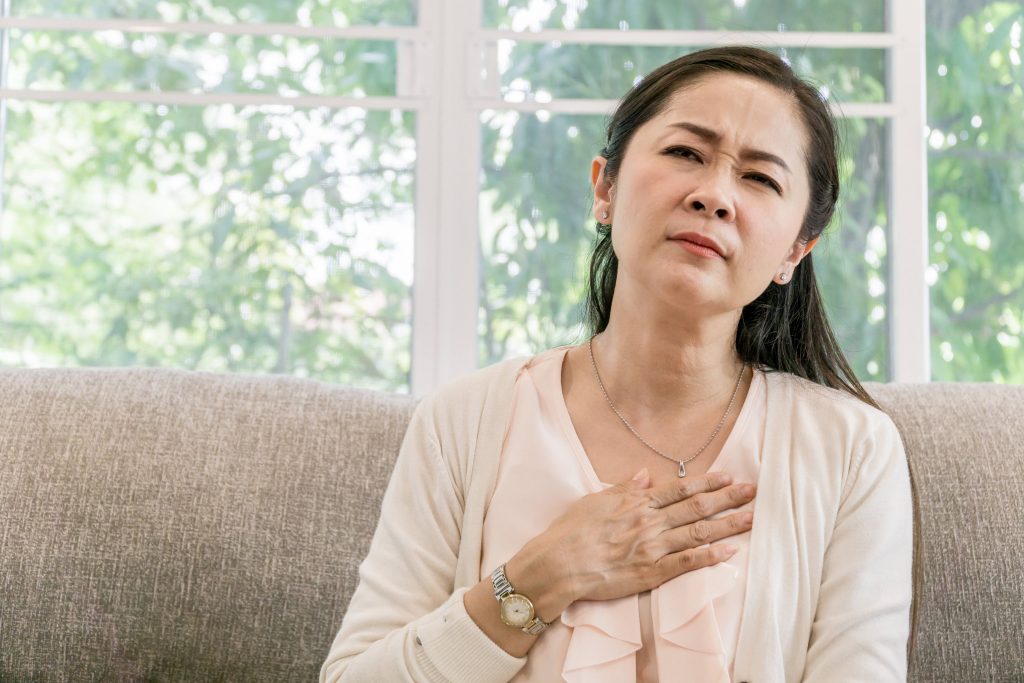 a middle aged Asian woman sitting on a couch near a window with her hand on her chest looking worried, sad, perhaps in pain