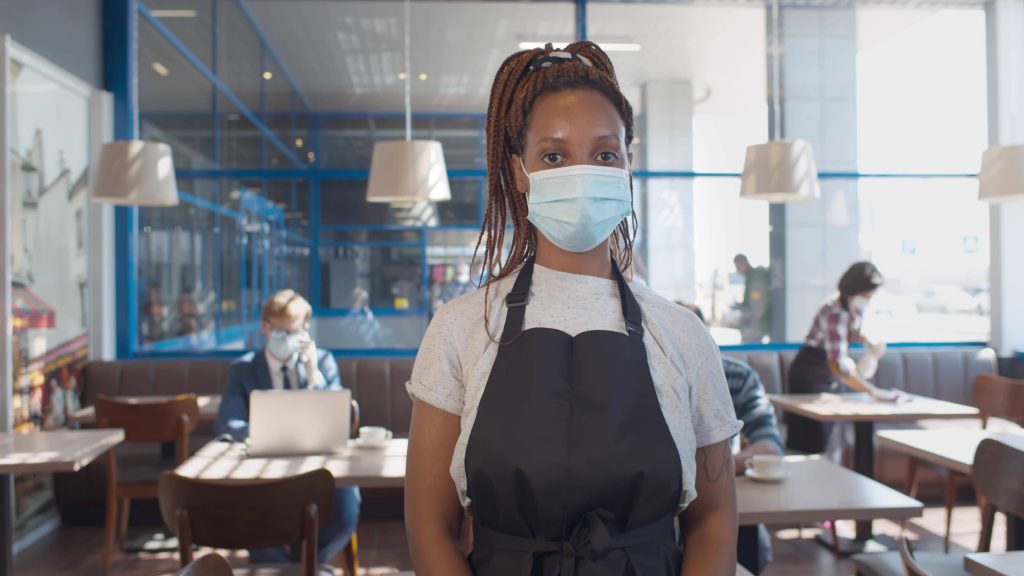 a young Black woman wearing a face mask and a work apron as a waitress in a restaurant