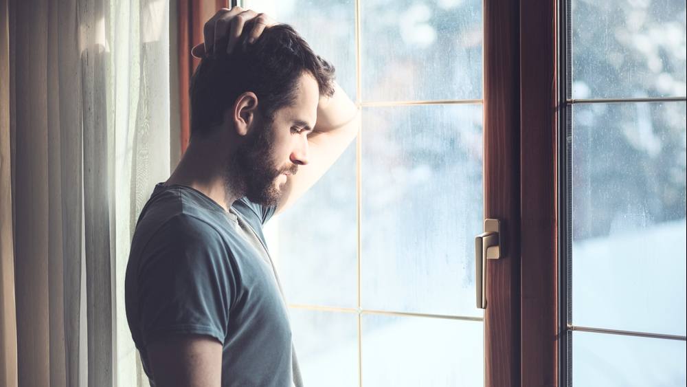 a young man standing by a rain-streaked window, looking sad, worried, depressed