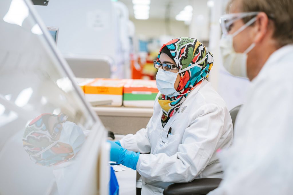 Mayo Clinic Laboratories researchers working on testing, a white man wearing glasses and a mask, and a white woman wearing glasses, gloves, a face mask and a hijab or scarf on her head