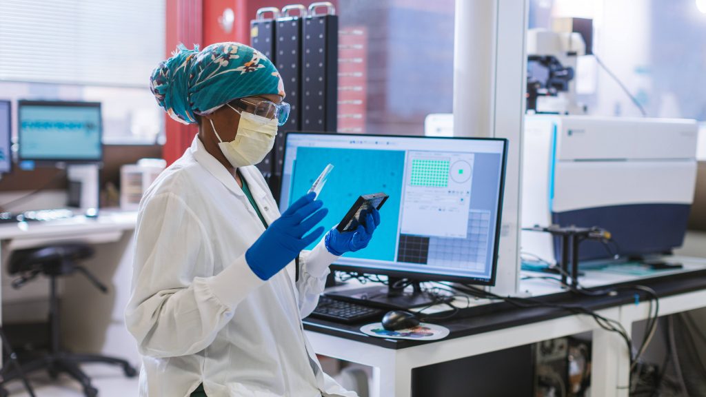 Mayo Clinic Laboratories health care researcher, a Black woman, wearing a hijab or scarf on her head and in PPE with a facemask, goggles and gloves looking at samples