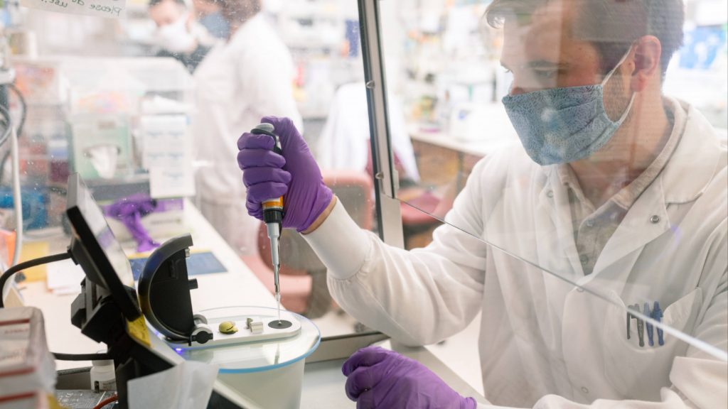 Mayo Clinic Laboratories researcher, a white man wearing a facemask and purple gloves while working with a specimen dropper behind a plastic shield in the lab