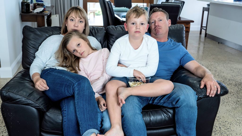 a family, a white woman and man sitting on a couch looking stressed, sad and tired, while holding their two white children on their laps