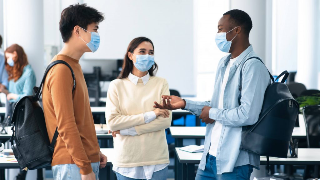 a group of three diverse college students, smiling and talking, wearing face masks and carrying backpacks in a school classroom