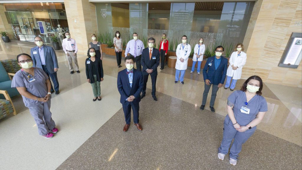 a large group of diverse people, Mayo Clinic medical staff members standing in a lobby wearing facemasks and practicing social distancing