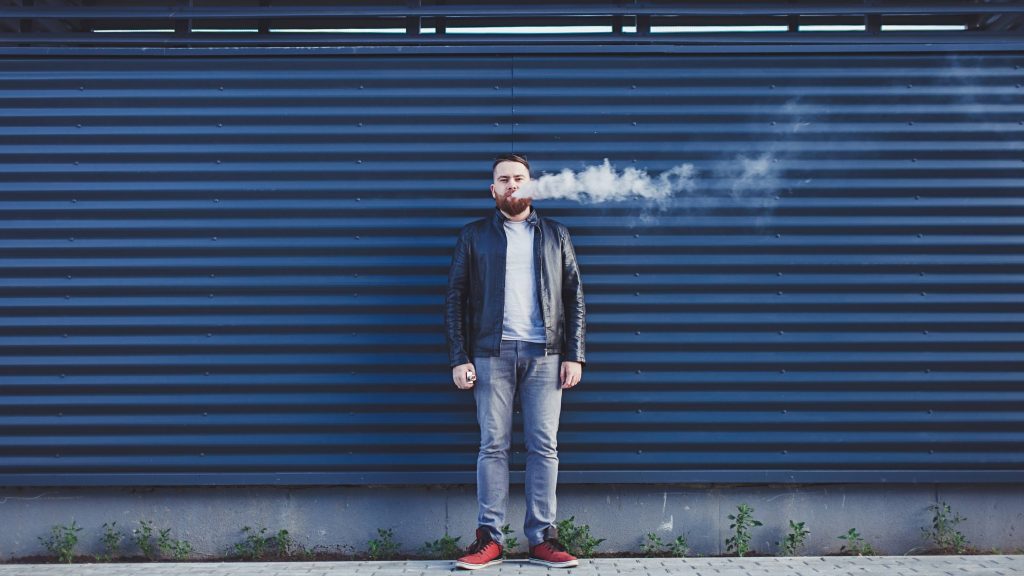 a white adult male in blue jeans standing outside in front of a blue metal building and smoking a cigarette or vaping