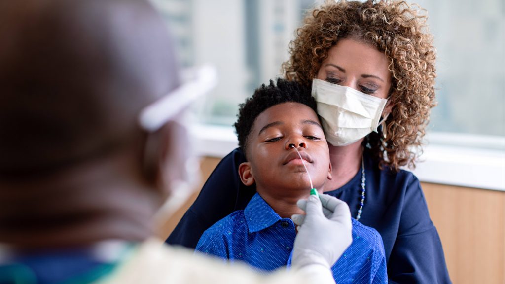 a white woman in hospital uniform and wearing a face mask, holding a young Black boy while a Mayo Clinic medical staff person performs a nasal COVID-19 test on the child