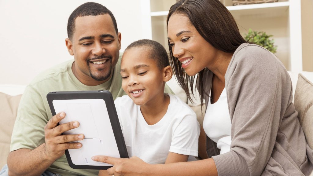 a young African American family, a Black man and Black woman, smiling and sitting on a couch with a young Black child looking at a computer tablet or iPad together