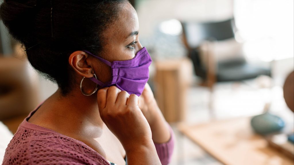 a young Black woman looking out a window and putting on a purple face mask