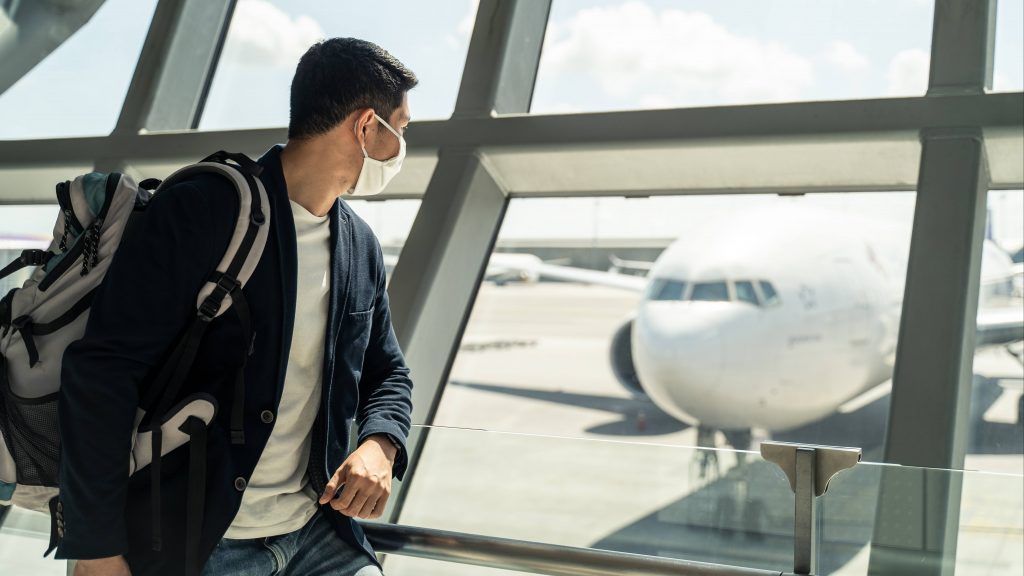 a young white, perhaps college aged man, wearing a back pack and a face mask, in an airport looking like he's ready to travel and watching an airplane out a window.