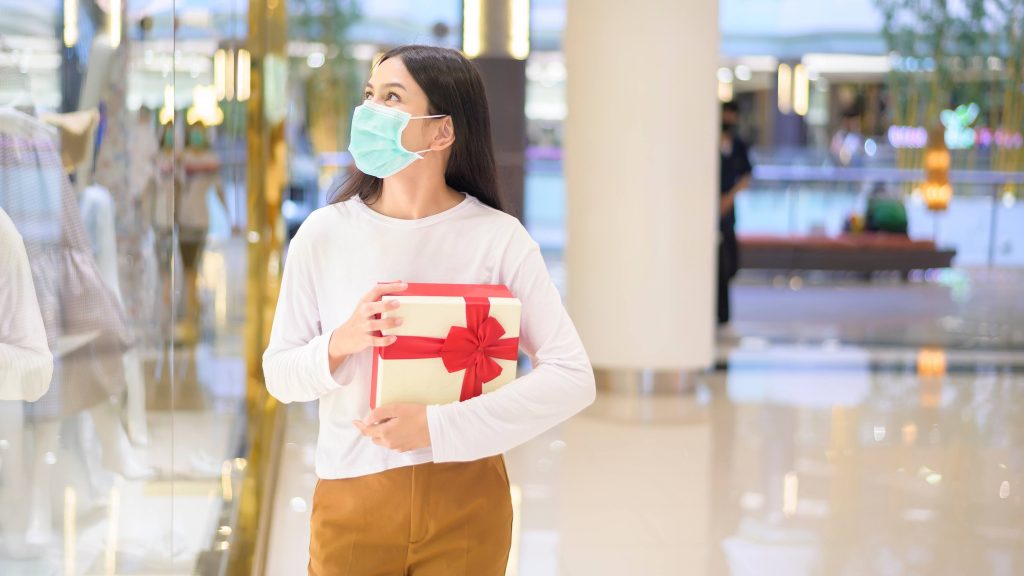 a young adult white woman shopping in a mall, wearing a face mask and carrying a gift box with a holiday bow