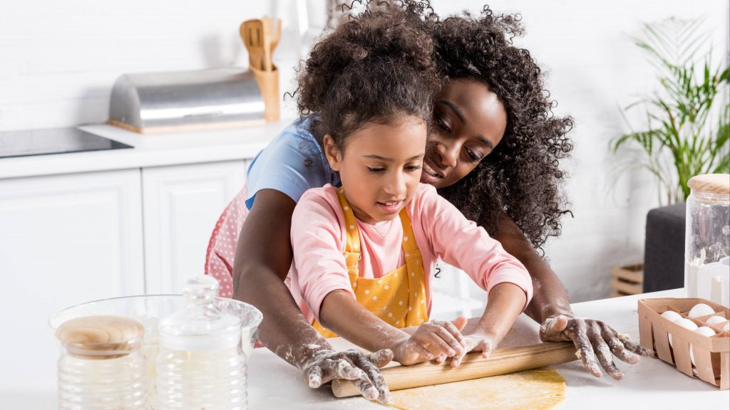 an adult Black woman, perhaps a mother, helping a little Black girl, perhaps her daughter, with baking in the kitchen and rolling out a pie crust