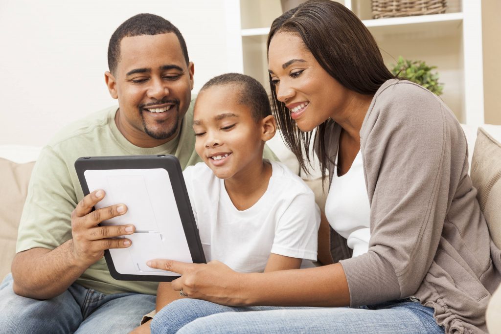 a young African American family, a Black man and Black woman, smiling and sitting on a couch with a young Black child looking at a computer tablet or iPad together