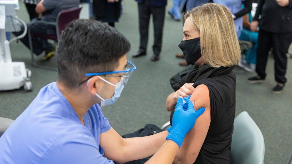 a Mayo Clinic in Florida employee, a white woman, getting her COVID-19 vaccine administered by a Mayo nurse , a man perhaps of Asian ethnicity wearing PPE