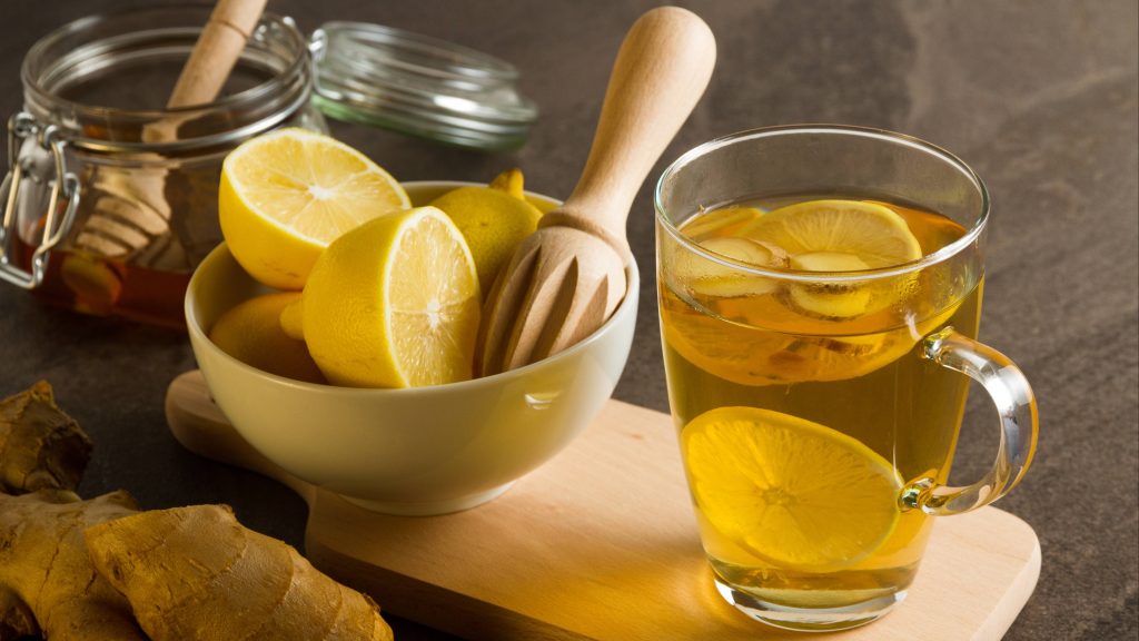 a kitchen counter with a bowl of lemons on a cutting board, a jar of honey, fresh ginger root and a clear glass of body cleansing juice