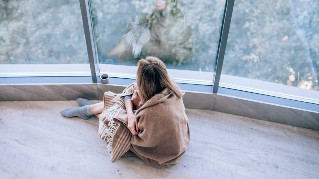 a lonely, sad looking white woman wrapped in a blanket and sitting near a window and looking out thoughtfully