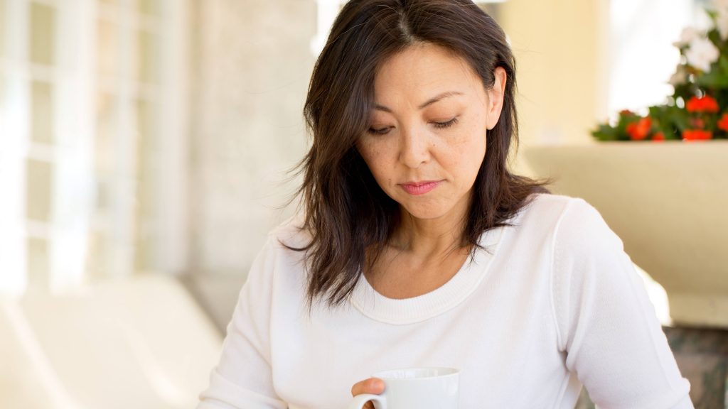 a middle aged woman, with light brown skin tone, perhaps Latina or Asian, holding a coffee mug and looking sad, worried, depressed