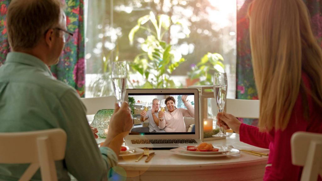a white man and a white woman sitting at a dinner table with glasses of champagne greeting friends or family members over a virtual meeting on a laptop