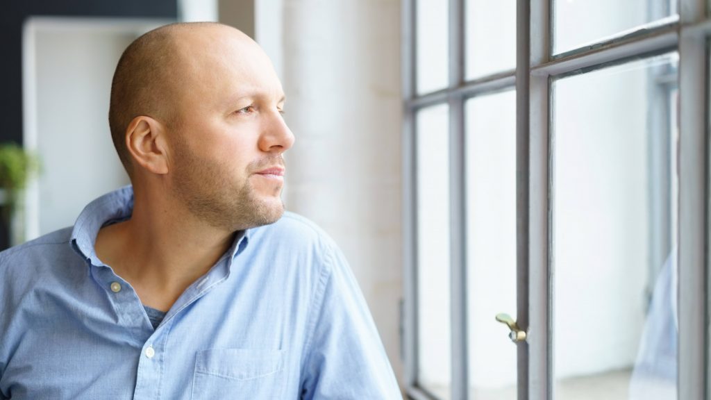 a white man in a blue shirt, looking serious, worried and thoughtful while standing at a window looking out