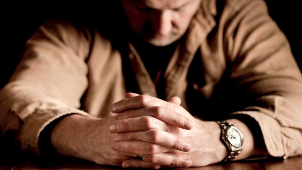 a white man with his hands clasped as if praying, sitting at table sad, thoughtful, depressed